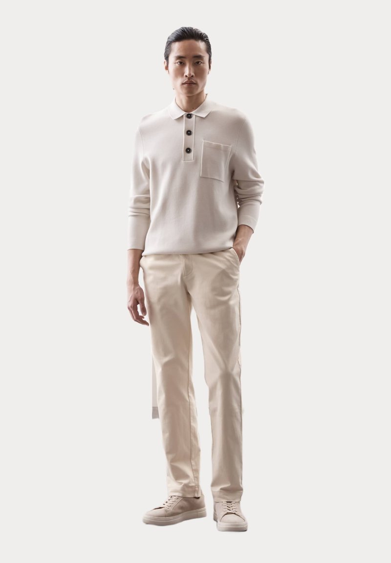 Lichtbeige gebreide polo met een kraag, drie donkere knopen en een borstzakje. Gepaard met lichtcrème broeken en beige sneakers.
