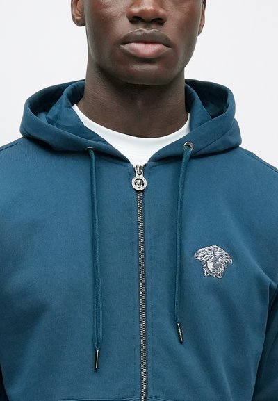 Tyrkysová mikina na zips s nastaviteľnými šnúrkami a výšivkou loga na prednej strane. Hladká látka s voľným výstrihom. Strieborný zips.