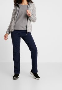 Veste zippée gris chevrons portée sur un haut gris, associée à un pantalon bleu marine. Chaussures noires à semelles blanches. Textures douces, design décontracté.