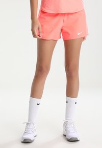 Pantalones cortos de mujer de Nike en un vibrante color rosa, hechos de un tejido ligero. Combinados con calcetines blancos hasta el tobillo que presentan un logo negro y zapatillas deportivas blancas.