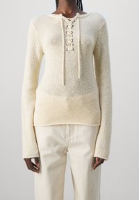 Pull en tricot crème à coupe décontractée, avec un col lacé, des manches longues et une finition texturée, associé à un pantalon blanc cassé.