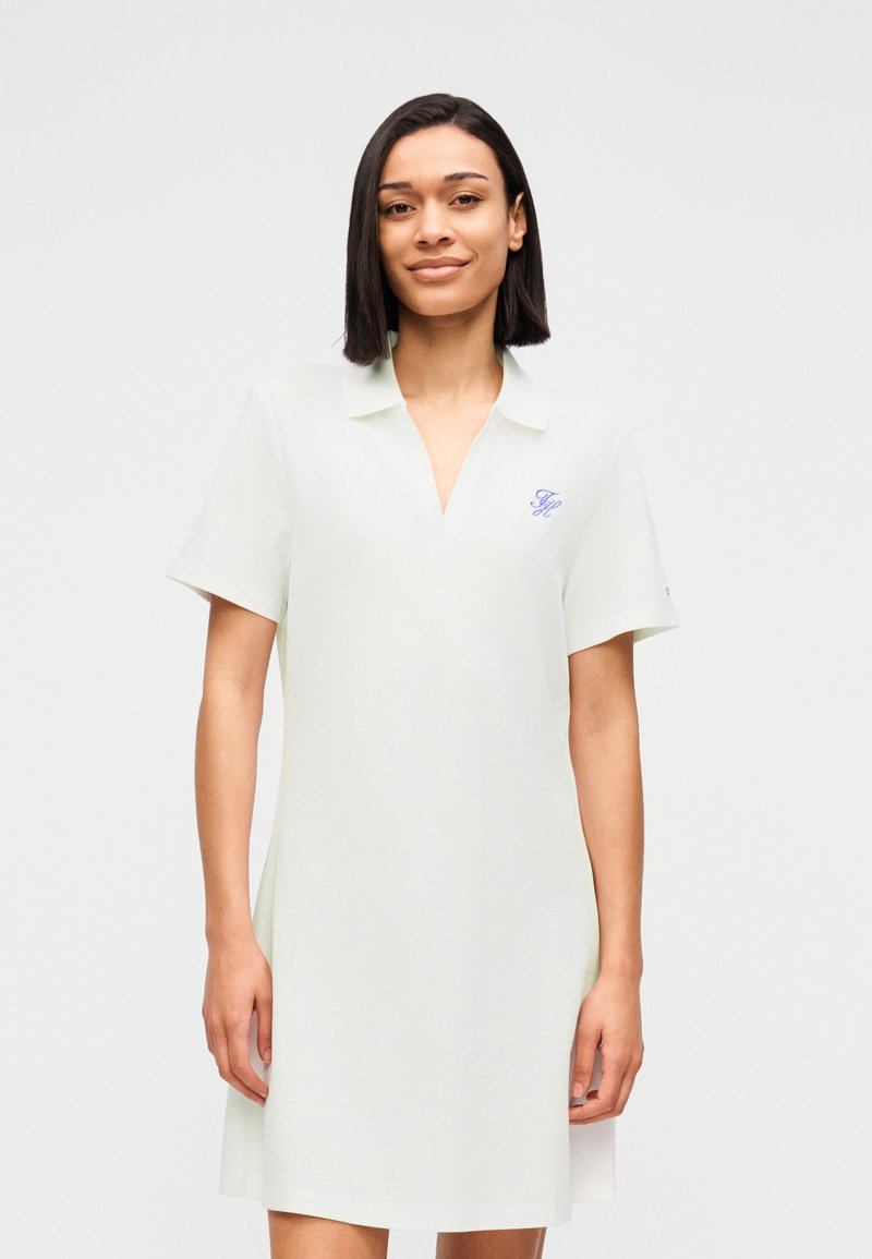 Jeune femme aux cheveux lisses et foncés portant une robe blanche à manches courtes avec col et un petit logo bleu sur la poitrine.