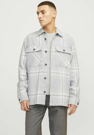 Camisa de franela a cuadros grises con dos bolsillos delanteros, botones negros y cuellorecto. Llevada sobre un suéter gris con un acabado texturizado.
