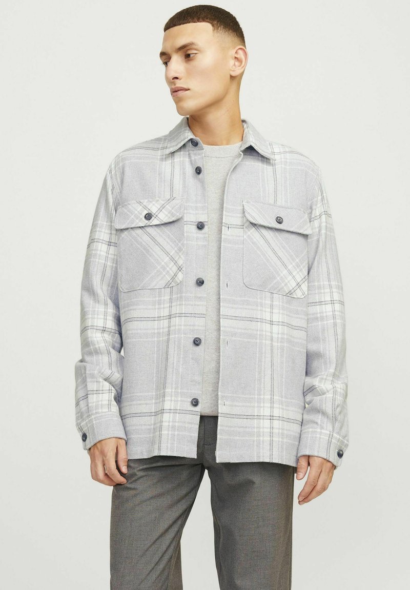 Camisa de franela a cuadros grises con dos bolsillos delanteros, botones negros y cuellorecto. Llevada sobre un suéter gris con un acabado texturizado.