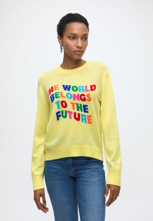 Kobieta ubrana w żółty sweter z kolorowym napisem „ŚWIAT NALEŻY DO PRZYSZŁOŚCI” i niebieskie jeansy, stojąca na jednolitym białym tle.