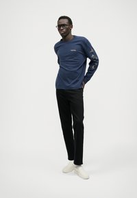 Tumesinine pika varrukaga särk, millel on rinna peal logo ja dekoratiivsed linnud varrukatel, koos mustade pükste ja valgete kingadega, stuudios. 