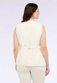 Chaleco sin mangas de color crema con un diseño ajustado y la parte posterior ceñida, que presenta un detalle de hebilla y cinta en la cintura. Textura suave.