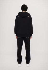 Černá mikina s velkou kapucí, volným střihem a rukávy s manžetami, doplněná černými tepláky. Minimalistický design, hladký povrch.