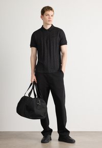 Melns polo krekls ar strukturētu rakstu, apvienots ar brīviem melniem biksēm. Modelis tur lielu melnu ceļojuma somu ar dubultām rokturiem.