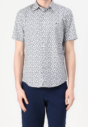 Man wearing a white short-sleeve button-up shirt with a small blue palm tree pattern and navy blue pants against a white background.