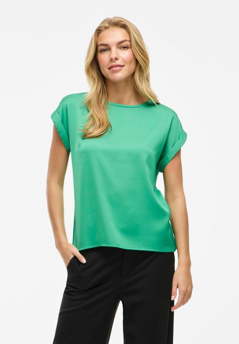 Blusa verde a maniche corte con scollo rotondo, texture liscia e maniche arrotolate, abbinata a pantaloni neri. Design semplice e moderno.