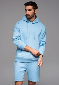 Ljusblå sweatshirt med huva och dragsnören, matchat med korta byxor; båda gjorda av mjukt, strukturerat tyg. Röd accentlogotyp på byxorna.