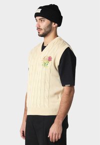 Krémová vesta se zaplétaným vzorem a výstřihem do V, zdobená červenou a žlutou květinovou výšivkou. Nositá přes černé tričko, doplněná černou čepicí.