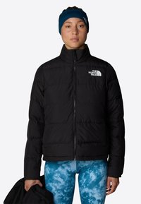 Czarna pikowana kurtka puchowa z zamkiem z przodu. Modelka ma na sobie niebieskie legginsy w wzory i trzyma inną kurtkę.