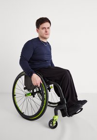 Egy fekete keretű, zöld kiegészítőkkel rendelkező kerekesszék. A felhasználó sötétkék hosszú ujjú pólót és fekete nadrágot visel, ülve, az egyik kezével a keréken.