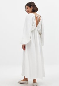 Weißes langärmliges Kleid aus strukturiertem Stoff, mit offenem Rücken und gebundenem Verschluss. Lässiges, luftiges Design, das bis zur Wadenmitte reicht, kombiniert mit leichter Fußbekleidung.