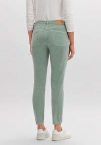 Jeans de pana verdes con corte slim, que cuentan con dos bolsillos traseros y un parche de marca marrón en la cintura, combinados con zapatillas deportivas blancas.