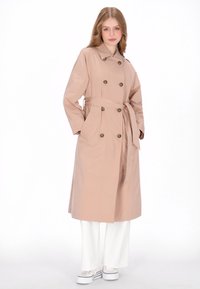 Lange beige trenchcoat met dubbele rij knopen, tailleceintuur en zijzakken. Gedragen over witte wijde broekspijpen en sneakers.