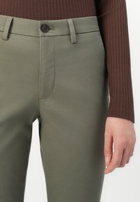 Olive green pants with a smooth texture, featuring a front button closure, side pockets, and a tapered fit, paired with a brown ribbed top.