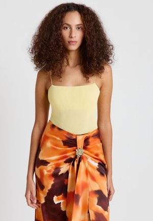 Femme aux cheveux bouclés portant un débardeur côtelé jaune pâle et une jupe à motifs orange, marron et noir avec une broche décorative.