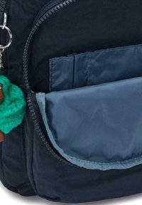 Zaino in tessuto blu navy con tasca anteriore con zip e interno blu, un charm verde morbido attaccato e una zip fluida.