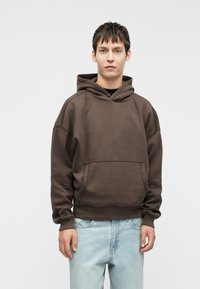 Felpa con cappuccio marrone in misto cotone, dotata di tasca a marsupio e polsini a coste, abbinata a jeans di denim blu chiaro.