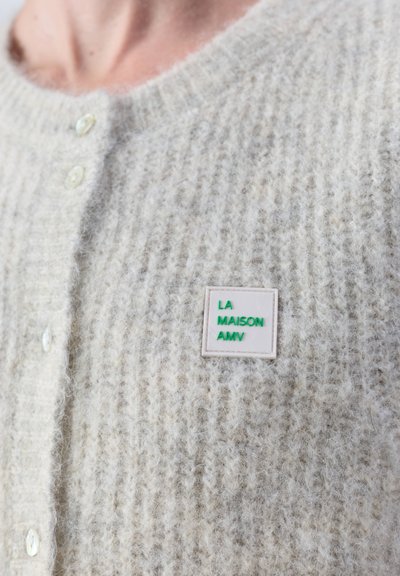 Cardigan en maille duveteuse gris clair avec des boutons en perle et une petite épingle carrée portant l'inscription "LA MAISON AMV" en lettres vertes.