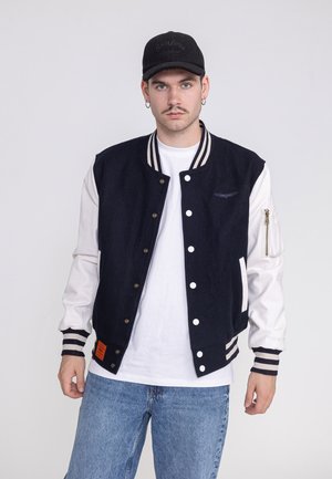 Junger Mann mit einer schwarz-weißen Varsity-Jacke, weißem Hemd, blauen Jeans und einer schwarzen Kappe, stehend vor einem schlichten hellen Hintergrund.