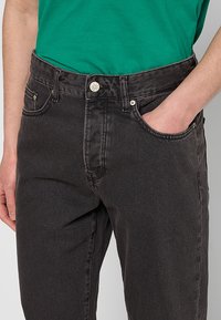 Jeans de mezclilla negra con un ligero desvanecimiento, diseño de cinco bolsillos, cierre de botón metálico y corte recto. Combinados con una camiseta verde.