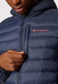 Námornícká pufovaná bunda s horizontálnym prešívaním, kapucňou a červeným logom "Champion" na hrudi. Obsahuje zipsovú kapsu pri ramene. Hladký povrch.