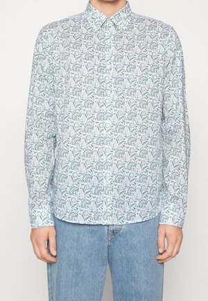 Mężczyzna ubrany w białą koszulę na guziki z zielonym kwiatowym wzorem i niebieskie dżinsy, stojący na tle jasnego, jednolitego tła.