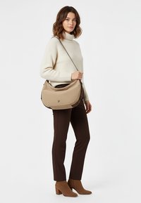 Naine kannab beeži kõrge kaelusega kampsunit, tumedat pruuni pükse, pruune pahkluu saabasteid ning kannab suurt beeži õlakoti.