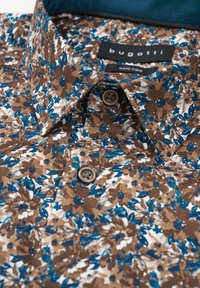 Braunes Blumenhemd mit blauen und weißen Akzenten. Mit einem Knopfkragen und zwei braunen Kunststoffknöpfen vorne. Aus weichem Baumwollstoff.