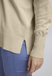 Beige kötött pulóver, bordás mandzsettával és alsó szegéllyel, oldalsó hasítékokkal. Világoskék nadrággal párosítva, sima textúrával és laza dizájnnal.