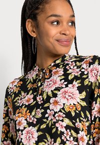 Blusa preta floral com flores rosa e laranja, detalhe de nó no pescoço e um tecido leve com uma textura suave.