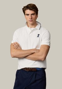 Jeune homme aux bras croisés portant un polo blanc avec le numéro 2 et un pantalon marine, sur un fond beige uni.
