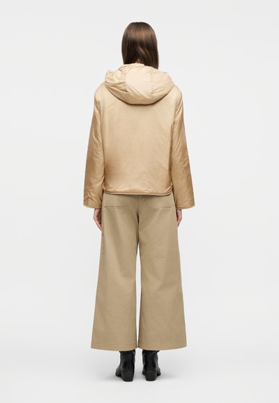 Άτομο με μπεζ μπουφάν με κουκούλα, παντελόνι χακί φαρδύ στους μηρούς και μαύρα μποτάκια μέχρι τον αστράγαλο, να στέκεται γυρισμένο με την πλάτη προς το απλό φόντο.