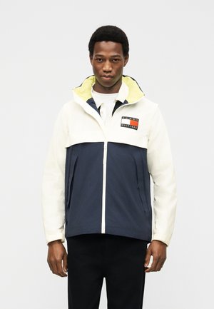 Férfi fehér és tengerészkék, blokkos mintájú dzsekiben, sárga kapucnibélésével és Tommy Hilfiger logóval a mellkasán, sima háttér előtt áll.