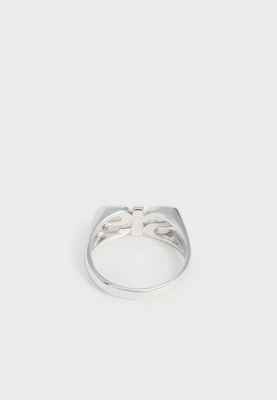 Bague en métal argenté avec une large bande et un design unique fendu sur le dessus, présentée sur un fond clair uni.