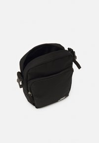 Borsa a tracolla in tessuto nero compatto con tasca frontale con zip, principale cerniera parzialmente aperta e cinturino laterale regolabile con fibbia.