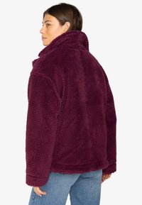 Cappotto in pelliccia sintetica bordeaux con colletto alto, esterno testurizzato e vestibilità rilassata, abbinato a jeans in denim blu.