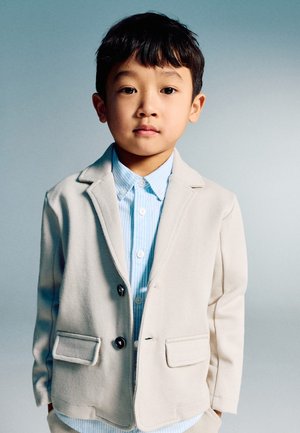 Junger Junge mit kurzen schwarzen Haaren, der einen beigen Blazer über einem hellblau gestreiften Hemd trägt und vor einem schlichten grauen Hintergrund steht.