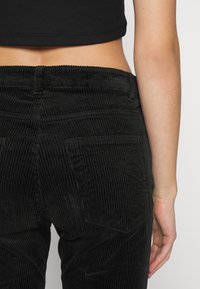 Zwarte ribfluwelen broek met verticale ribbels, een hoge taille en achterzakken, gezien vanuit de achterkant. Glad materiaal, nauwsluitende pasvorm.