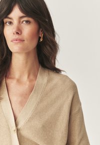 Femme aux cheveux bruns portant des boucles d'oreilles en or et un cardigan en tricot beige, sur fond clair uni, regardant vers l'avant.