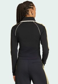 Černá sportovní bunda s dlouhými rukávy, s bílými a žlutými bočními pruhy, límečkem a přiléhavým střihem, vyrobená z elastického materiálu.