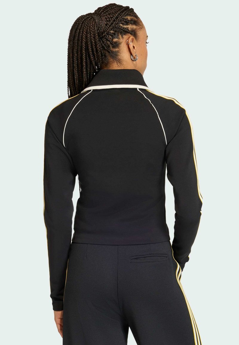 Sort atletisk jakke med lange ærmer, har hvide og gule striber på siden, krave og tætsiddende design, lavet af elastisk materiale.