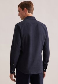 Uomo in piedi di spalle, vestito con una camicia a maniche lunghe blu scuro con bottoni e pantaloni scuri abbinati, su uno sfondo neutro.