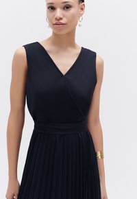 Abito a pieghe blu navy con scollatura a V, design senza maniche e vita con cintura. Caratterizzato da un tessuto testurizzato e da un accessorio a bracciale dorato scintillante.