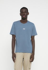 Camiseta azul de algodón con cuello redondo y mangas cortas. Incluye un pequeño logo blanco estampado en el pecho. Combinada con pantalones cargo beige.