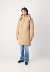 Beige lang pufferjakke med glidelås front, rosa hettefôr og sidelommer. Kombinert med lyseblå vide jeans og svarte sko.